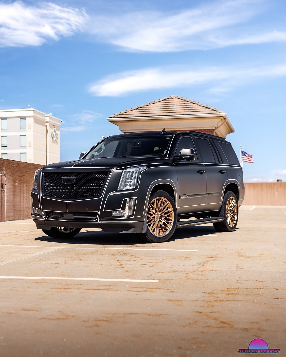 Black Cadillac Escalade SUV with bronze wheels parked on a rooftop, clear blue sky.