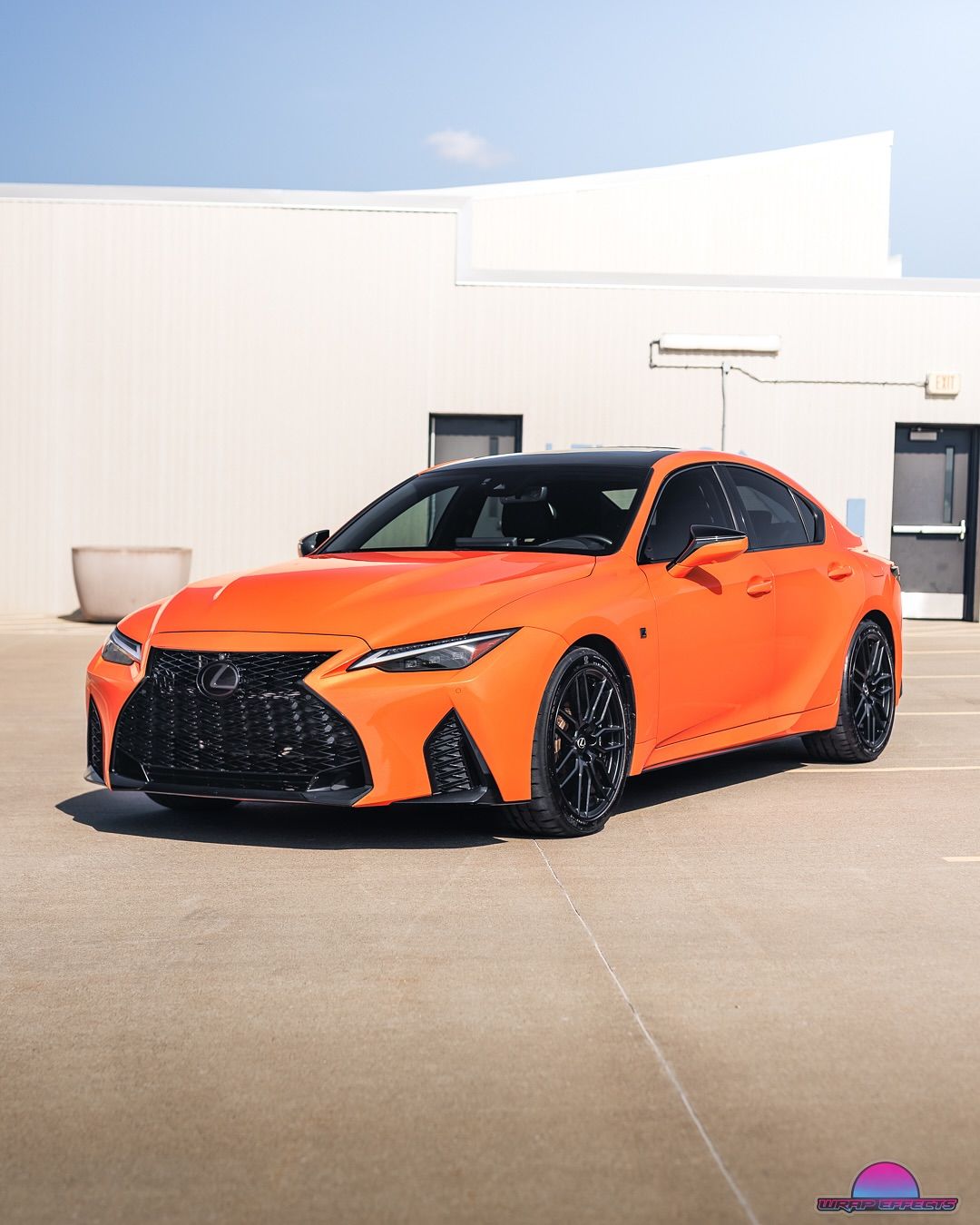 Orange Lexus sedan parked in front of a building, black wheels, sunny day.