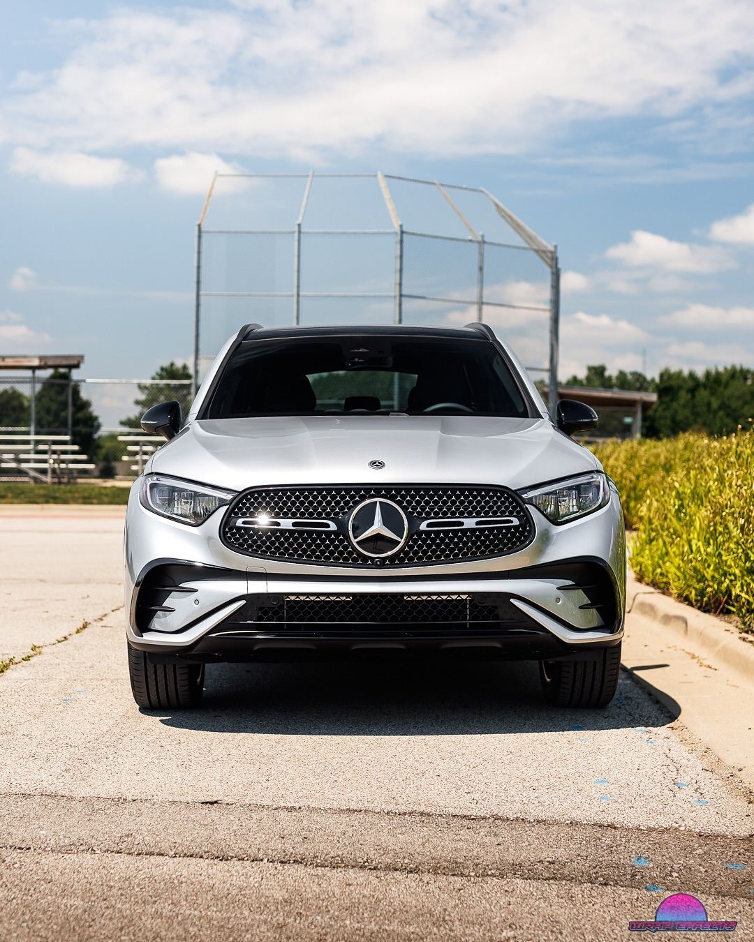 Silver Mercedes-Benz SUV parked on gravel, in front of a baseball field, with a blue sky in the background.