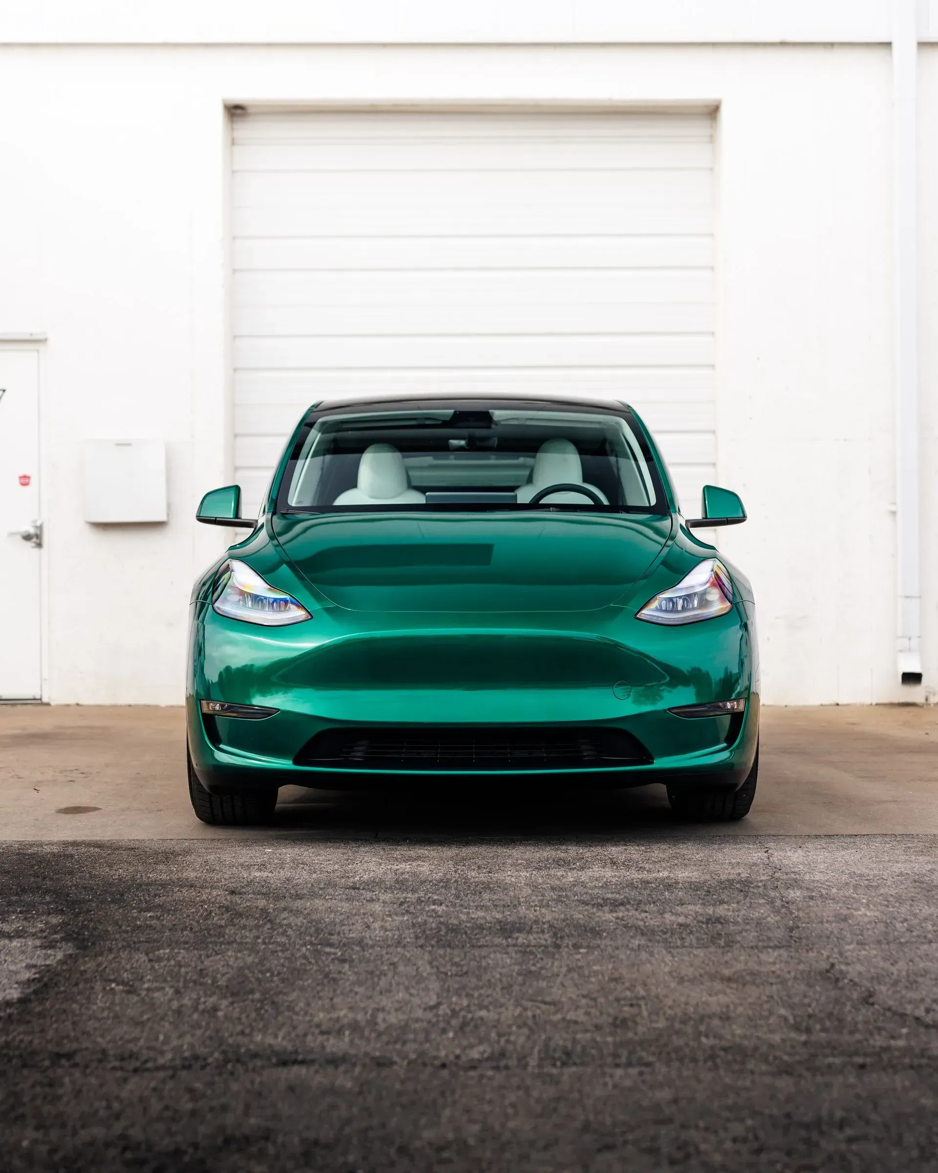 Green Tesla Model Y parked in front of a white building with a garage door.
