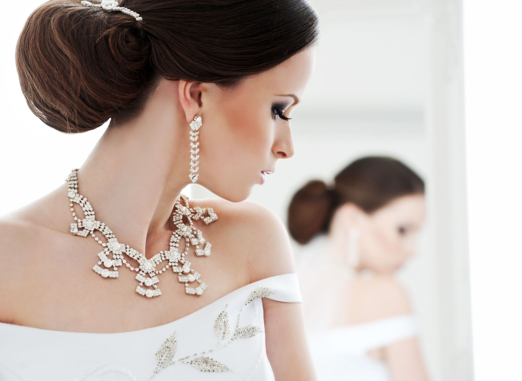 Bride in white dress, looking down with diamond necklace and earrings. Hair updo, mirror reflection.