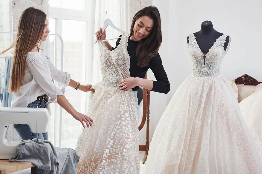 Two women examining a wedding dress in a bridal shop. One holds it up, the other observes.