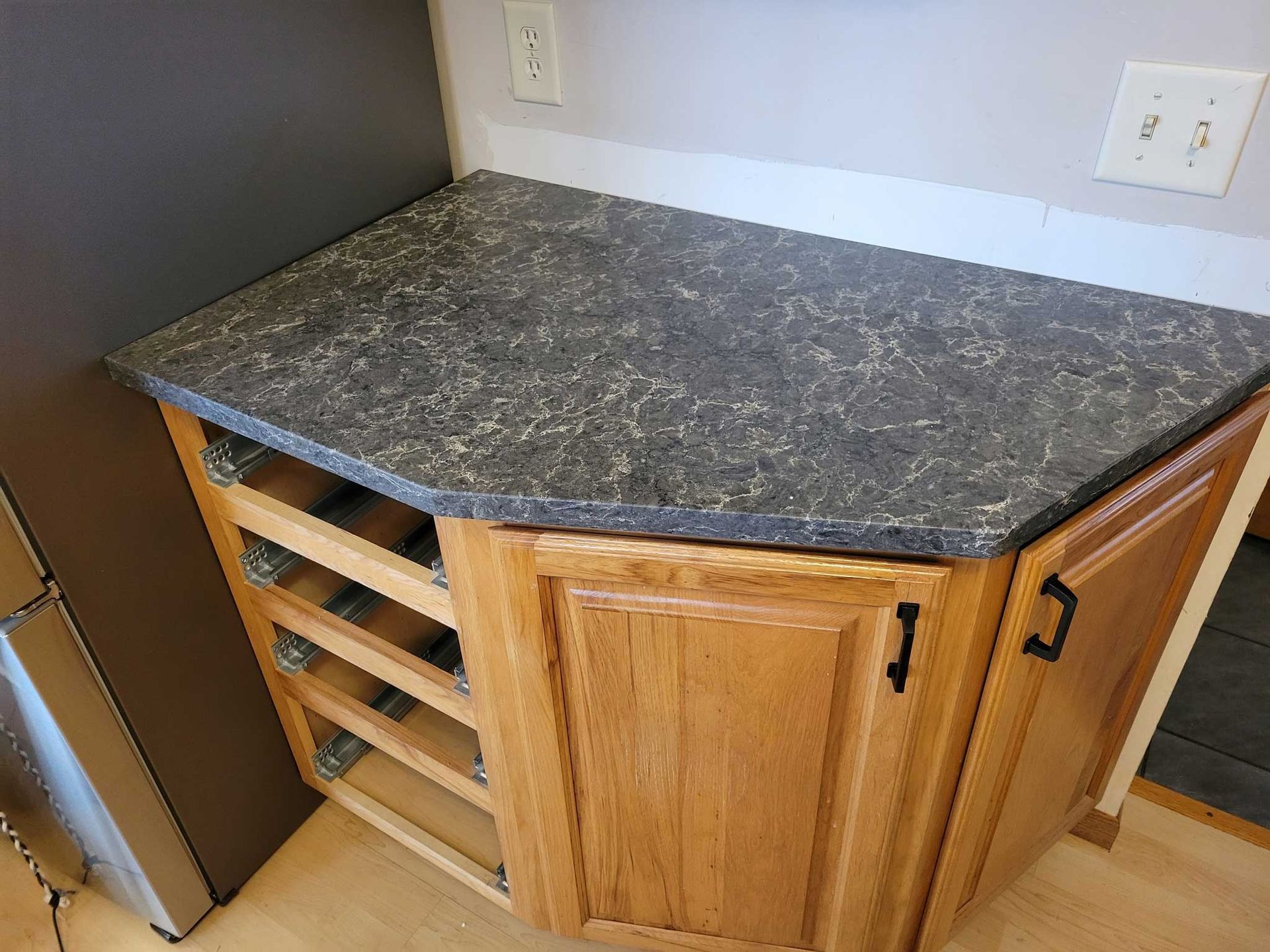 A kitchen cabinet with a dark countertop and open drawer slides; a refrigerator is on the left.