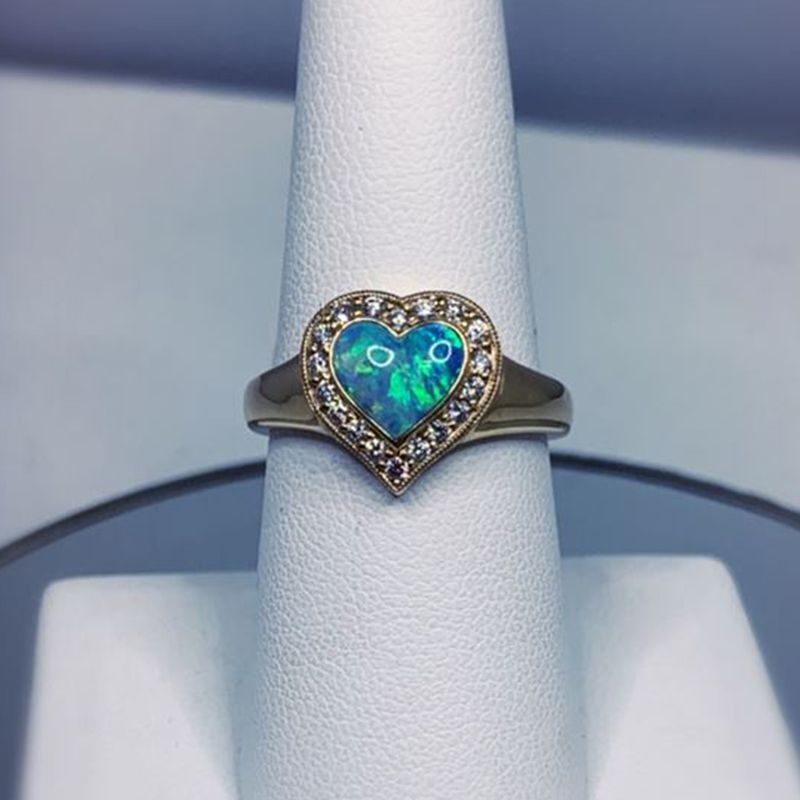 A ring with a heart shaped opal and diamonds on a white ring holder.