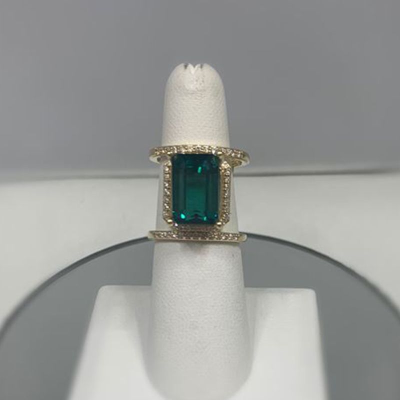 A ring with an emerald and diamonds is sitting on a white ring holder.