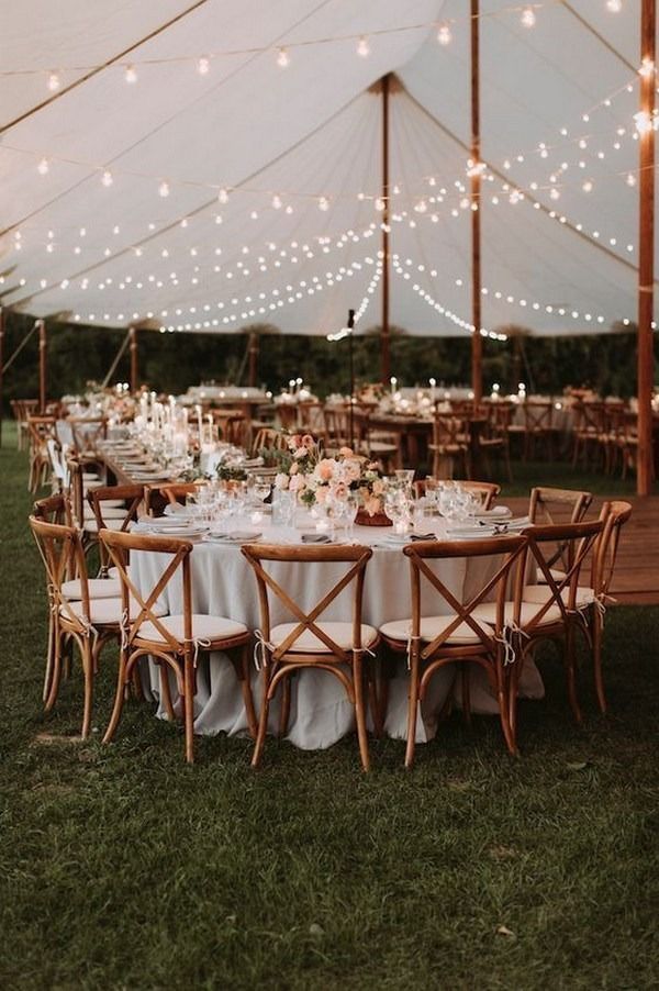 Round tables set under a white tent with string lights at an outdoor evening reception