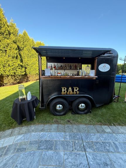 Black mobile bar trailer with serving window on a lawn, beside a small cocktail table and trees