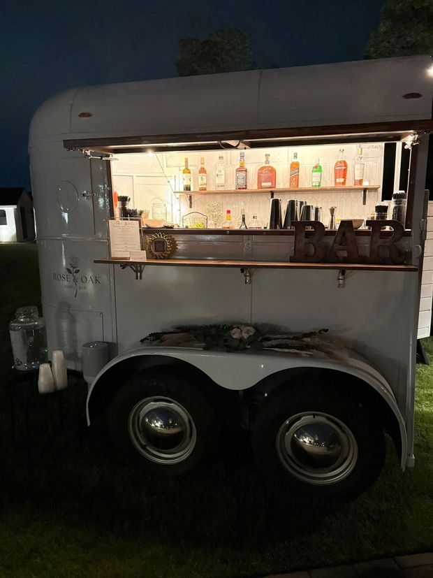 Small white trailer bar at night with a lit serving window and colorful bottles on display.