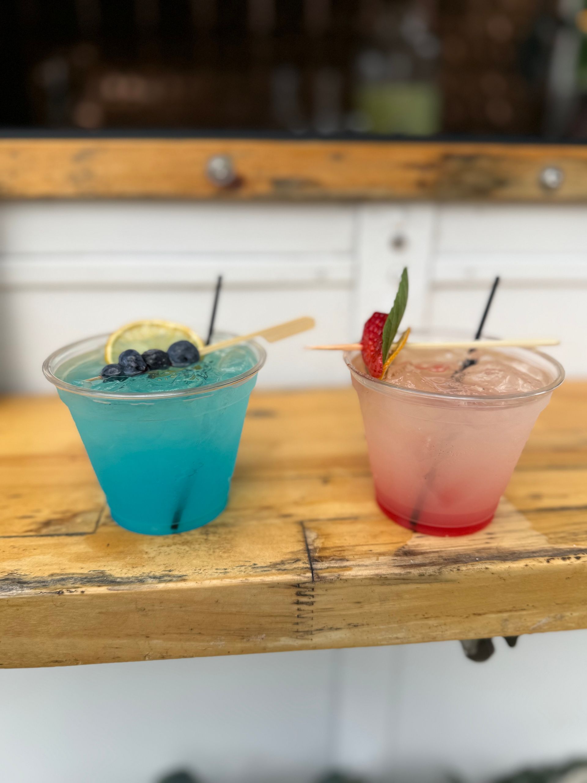 Two cocktails: blue with blueberries and a lemon slice, pink with strawberry and mint, on a wooden bar.