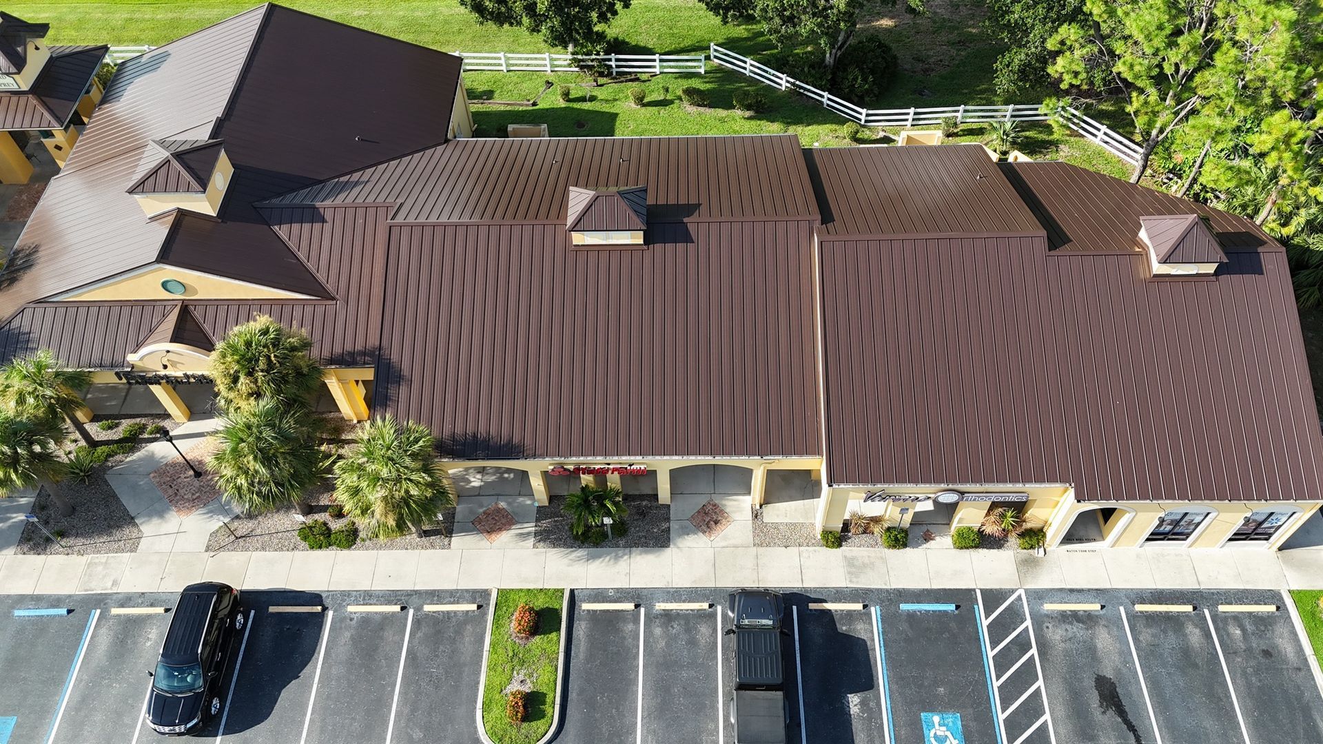 An aerial view of a building with a brown metal roof, with a parking lot in front and trees in the background.
