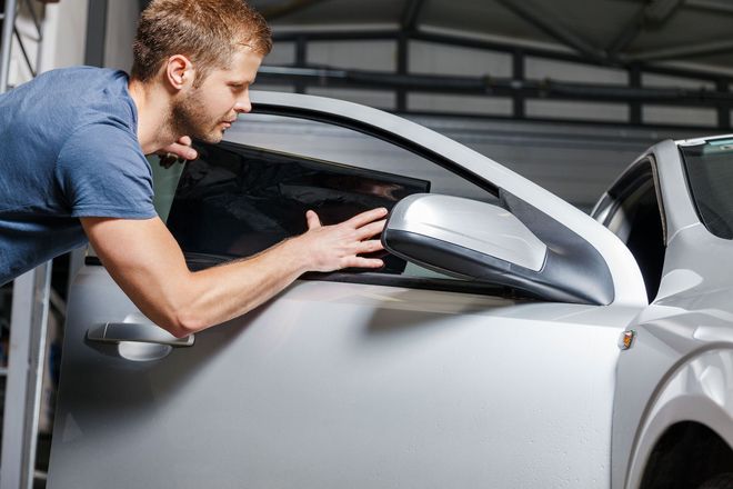 Man leaning into a silver car, inspecting or cleaning the side mirror in a garage.