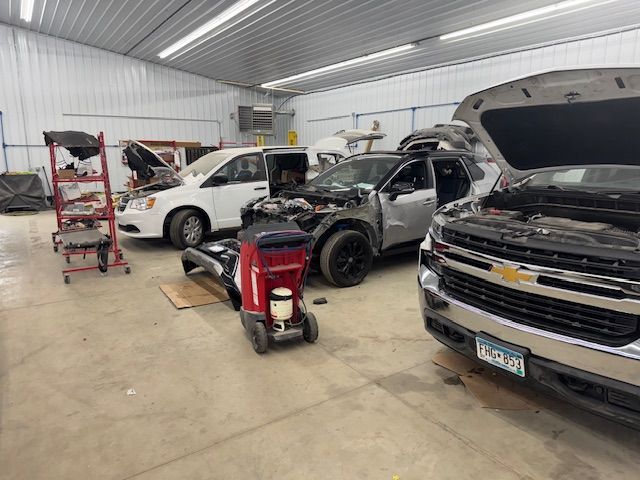 A garage with multiple vehicles undergoing repair; including a white van, gray SUV, and silver truck. Tools and equipment are scattered about.