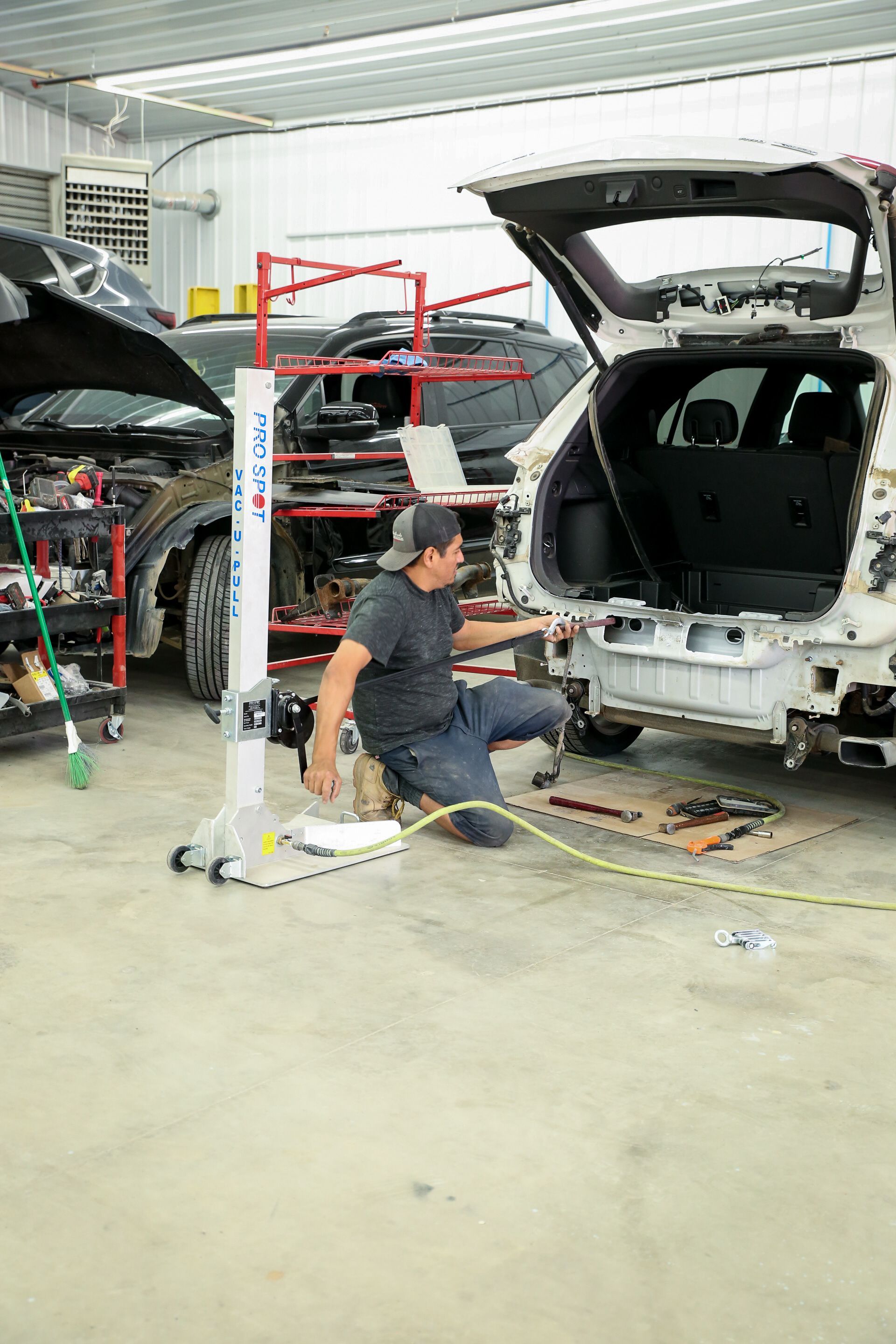 Man in auto body shop working on a car's rear panel section. He's kneeling by the bumper with tools and a vertical straightening machine nearby.