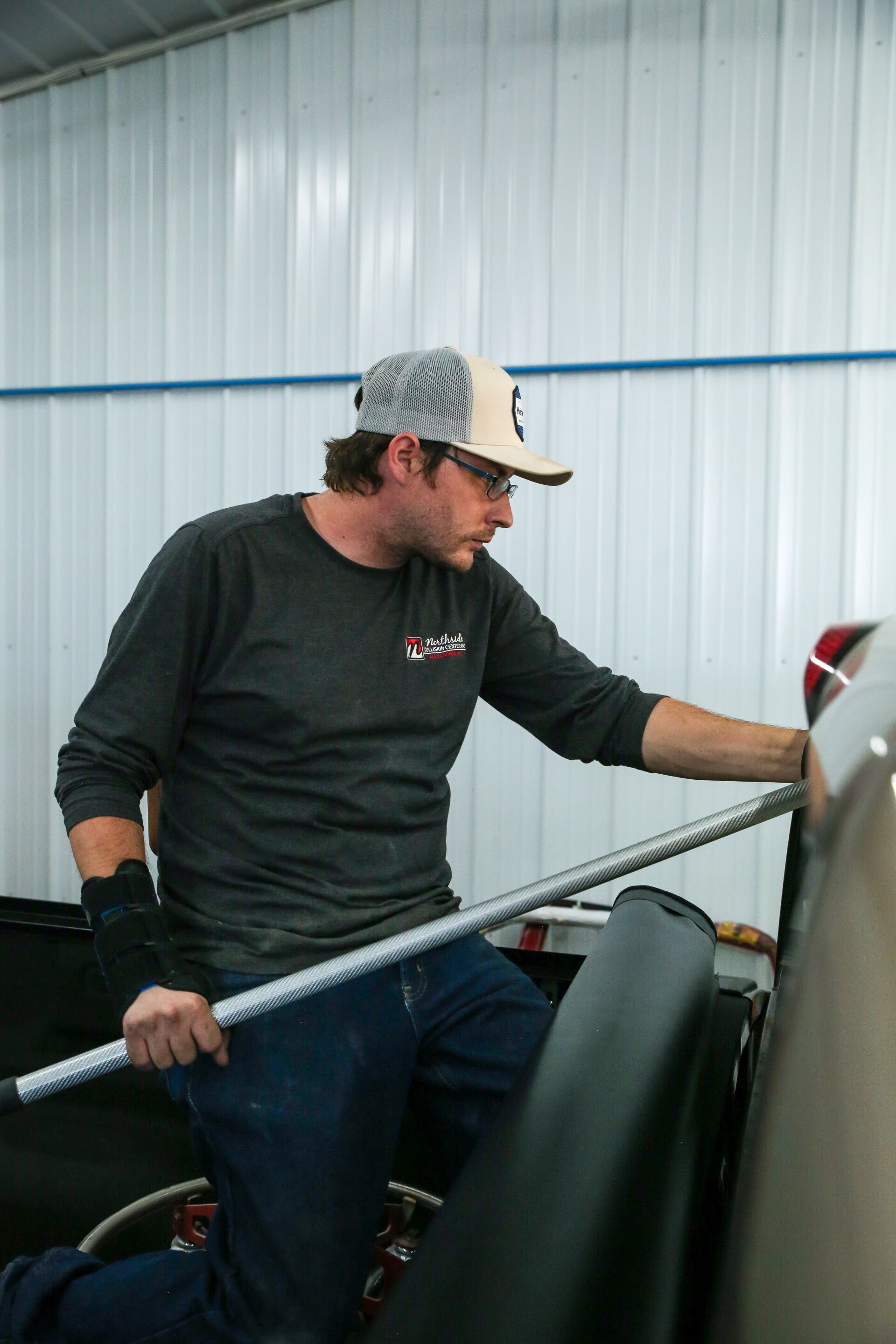 Man in a cap and glasses kneels, working on a car with a metal tool inside a garage.