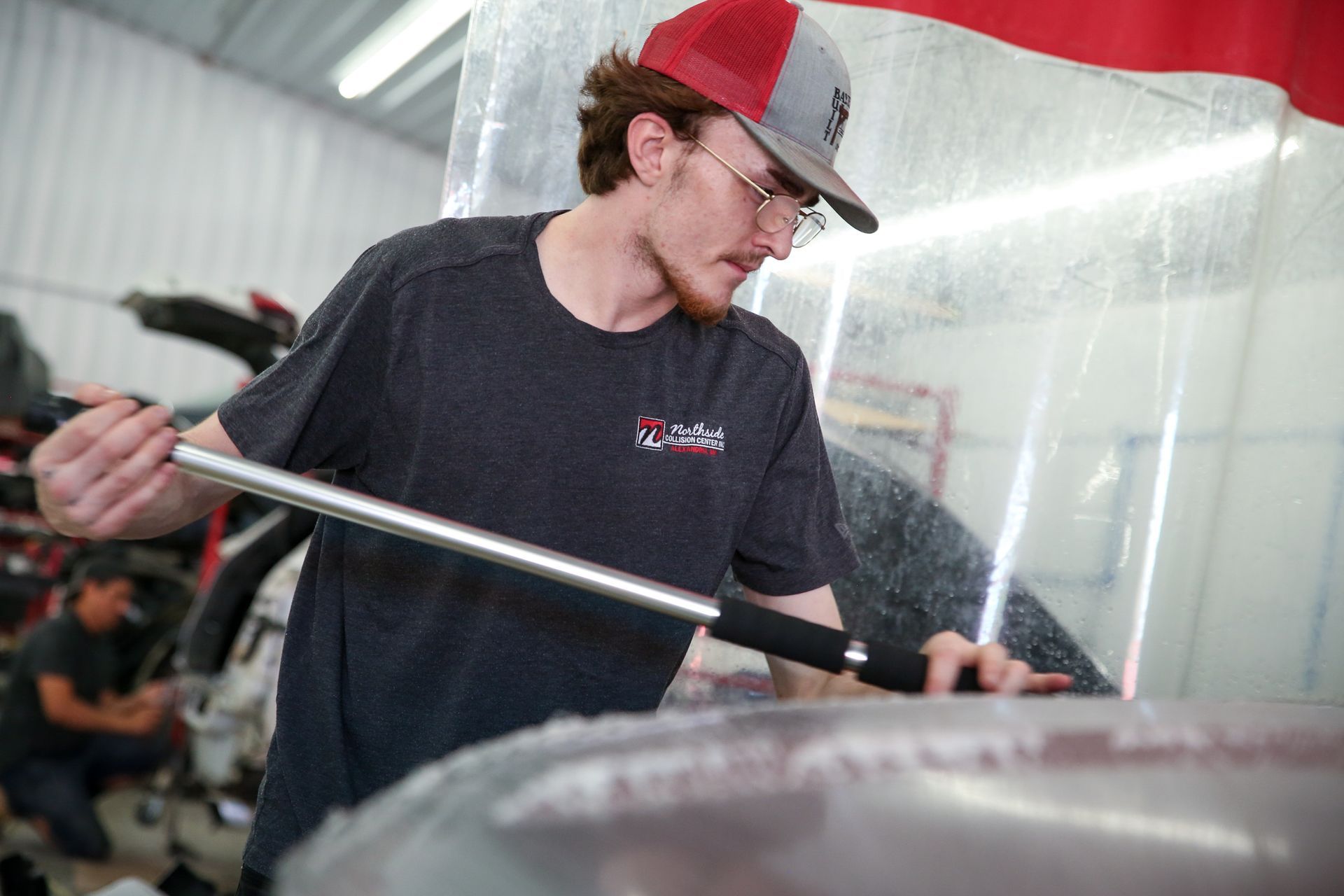 A auto body technician in a cap and glasses works on a car, holding a tool in an auto body shop.