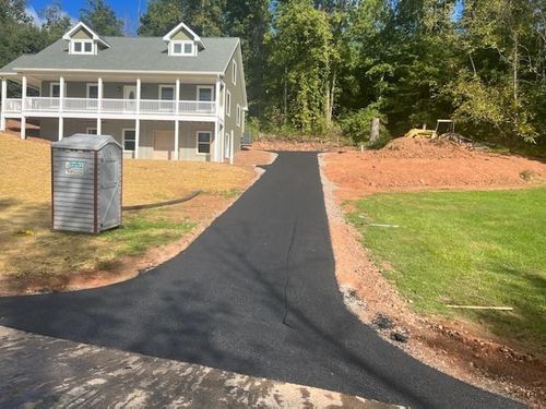Black driveway leading to a white house with a porch, surrounded by grass and trees.