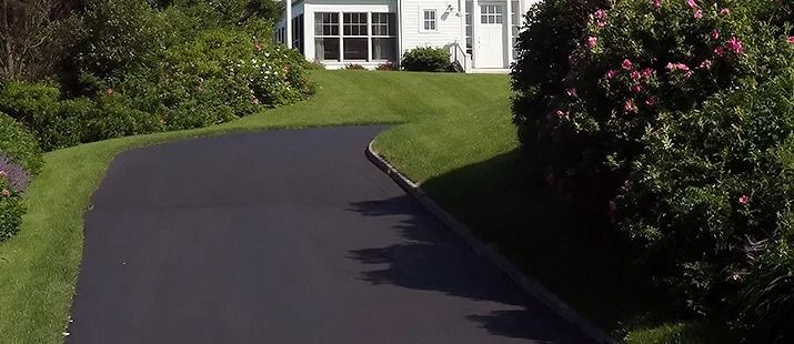 Freshly paved black driveway curving toward a white house with green lawn and shrubs