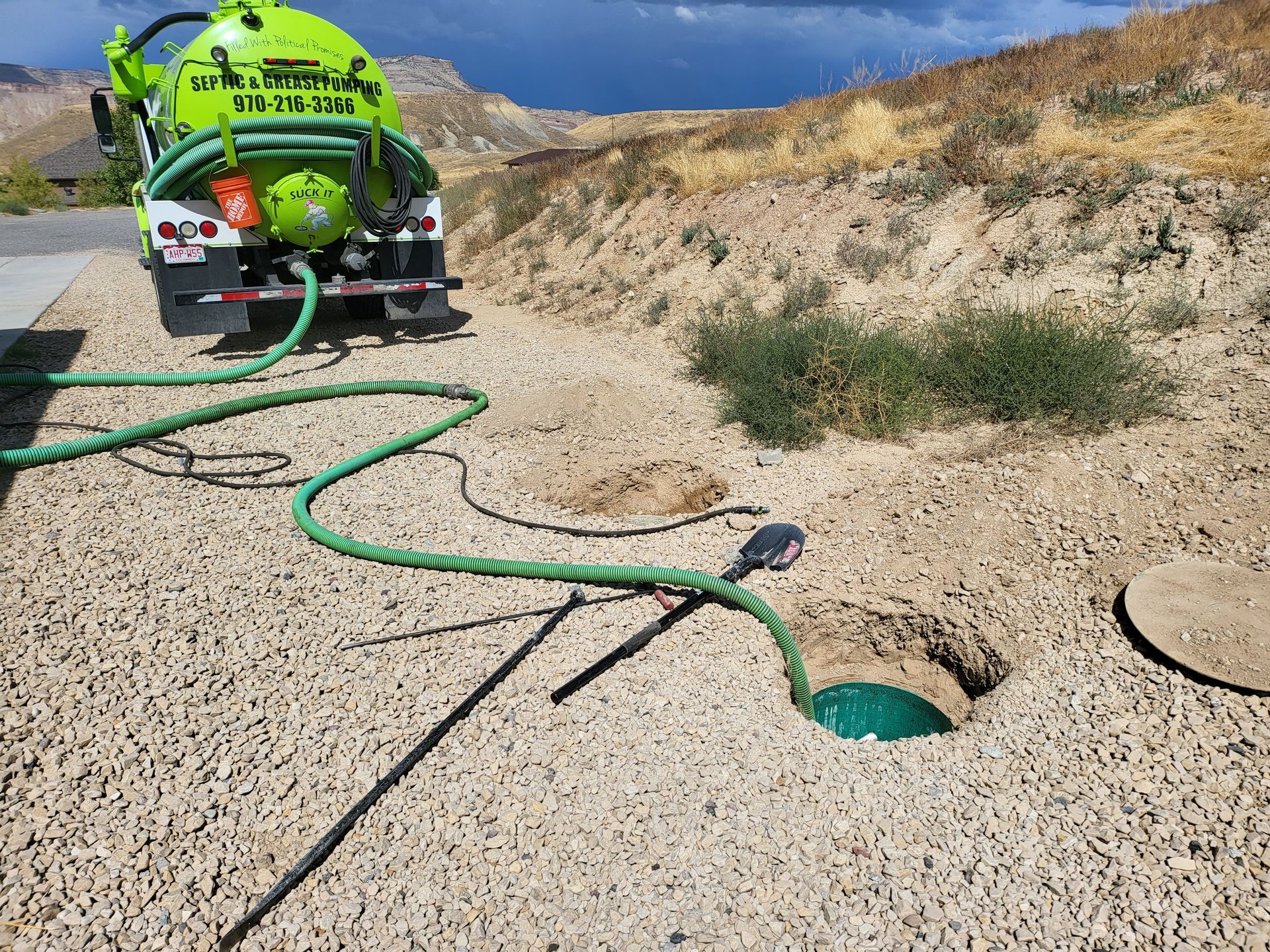 Bright green vacuum truck pumping from an open manhole in a sandy roadside ditch.