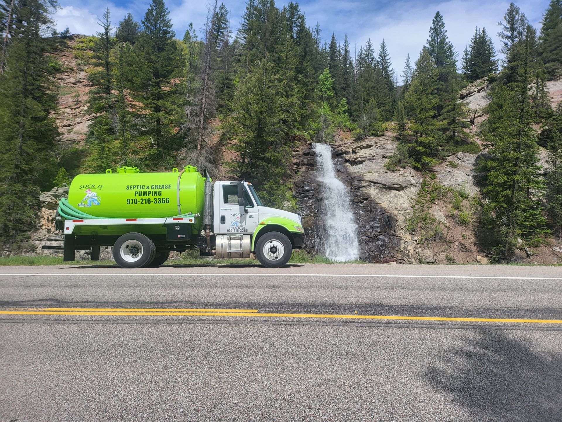 Green utility truck parked by a small waterfall on a rocky roadside in a pine forest