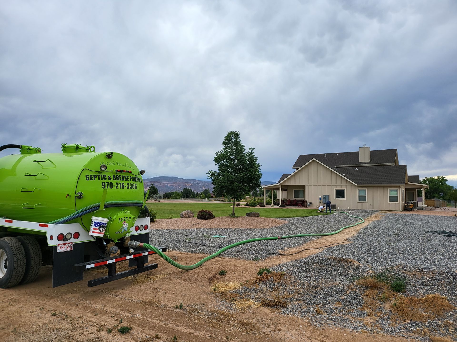 Green water tanker on a gravel lot beside a beige house under a cloudy sky