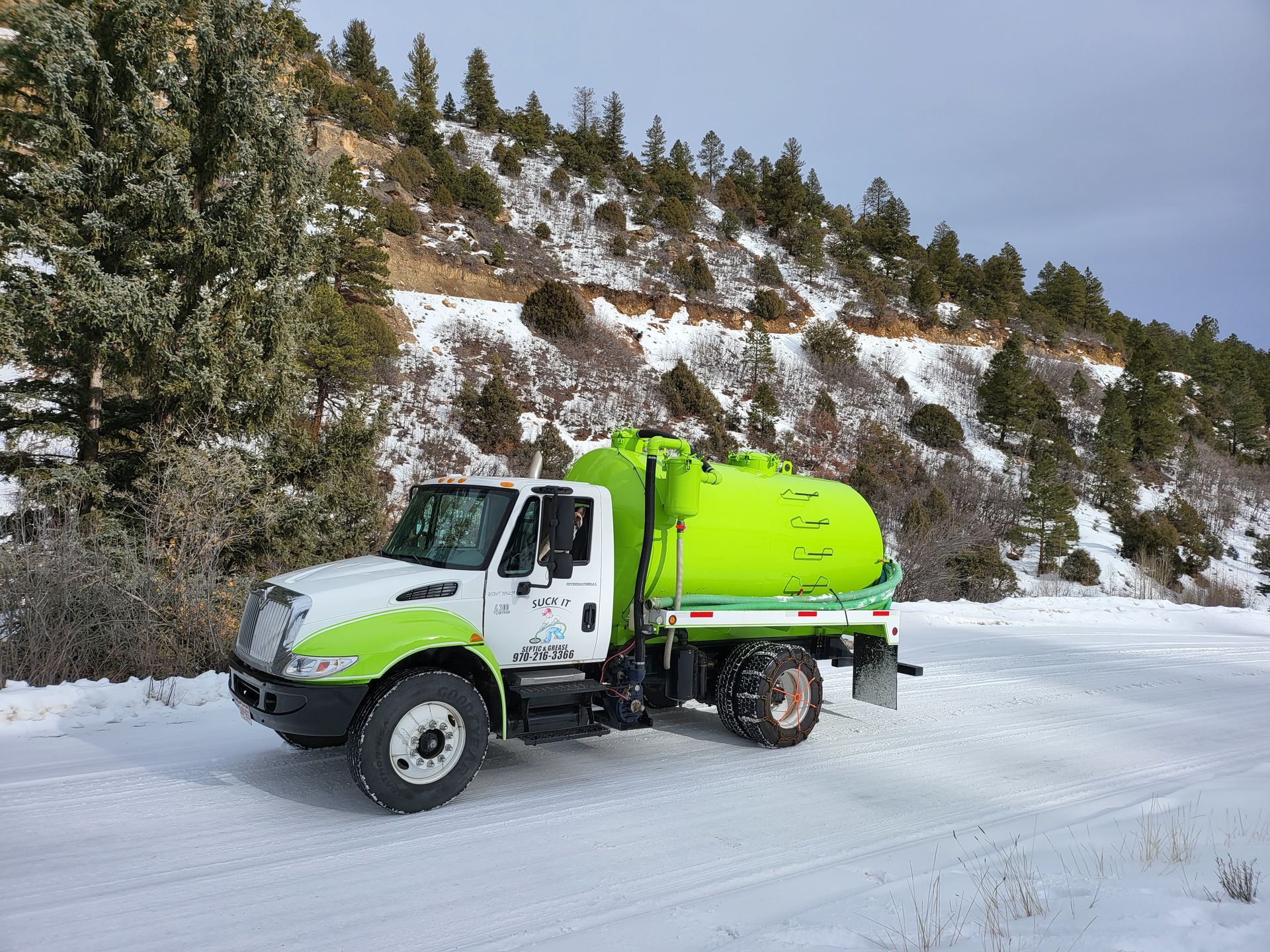 Bright green tanker truck driving on a snowy mountain road beside a hillside of trees