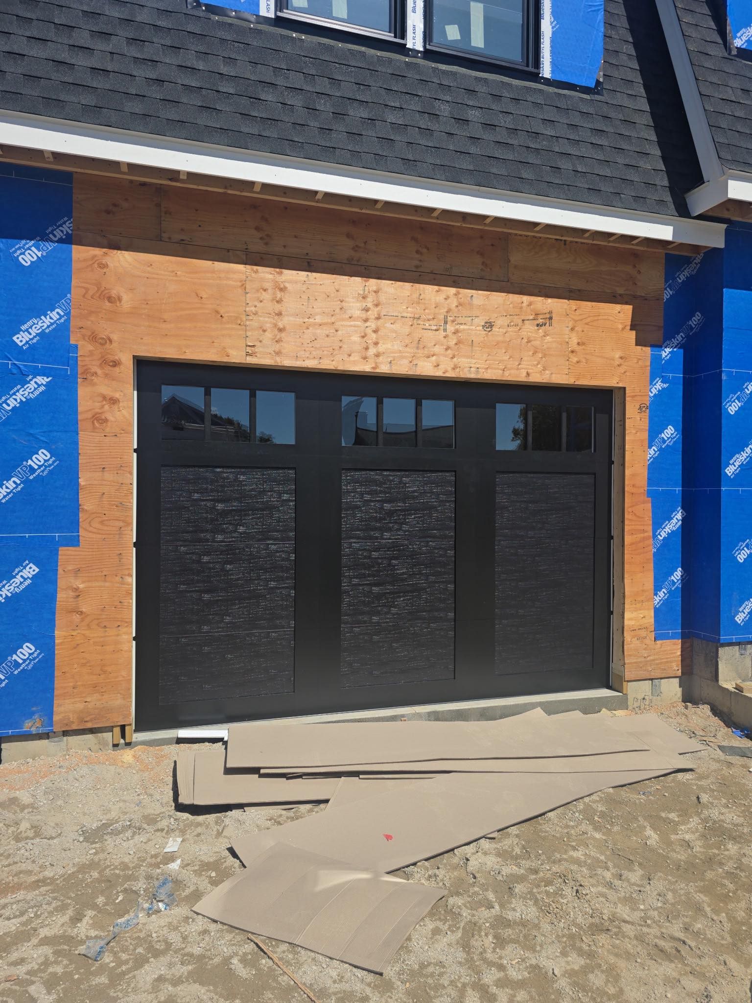 Black garage door under construction, blue paper siding, wood framing, and black roof.