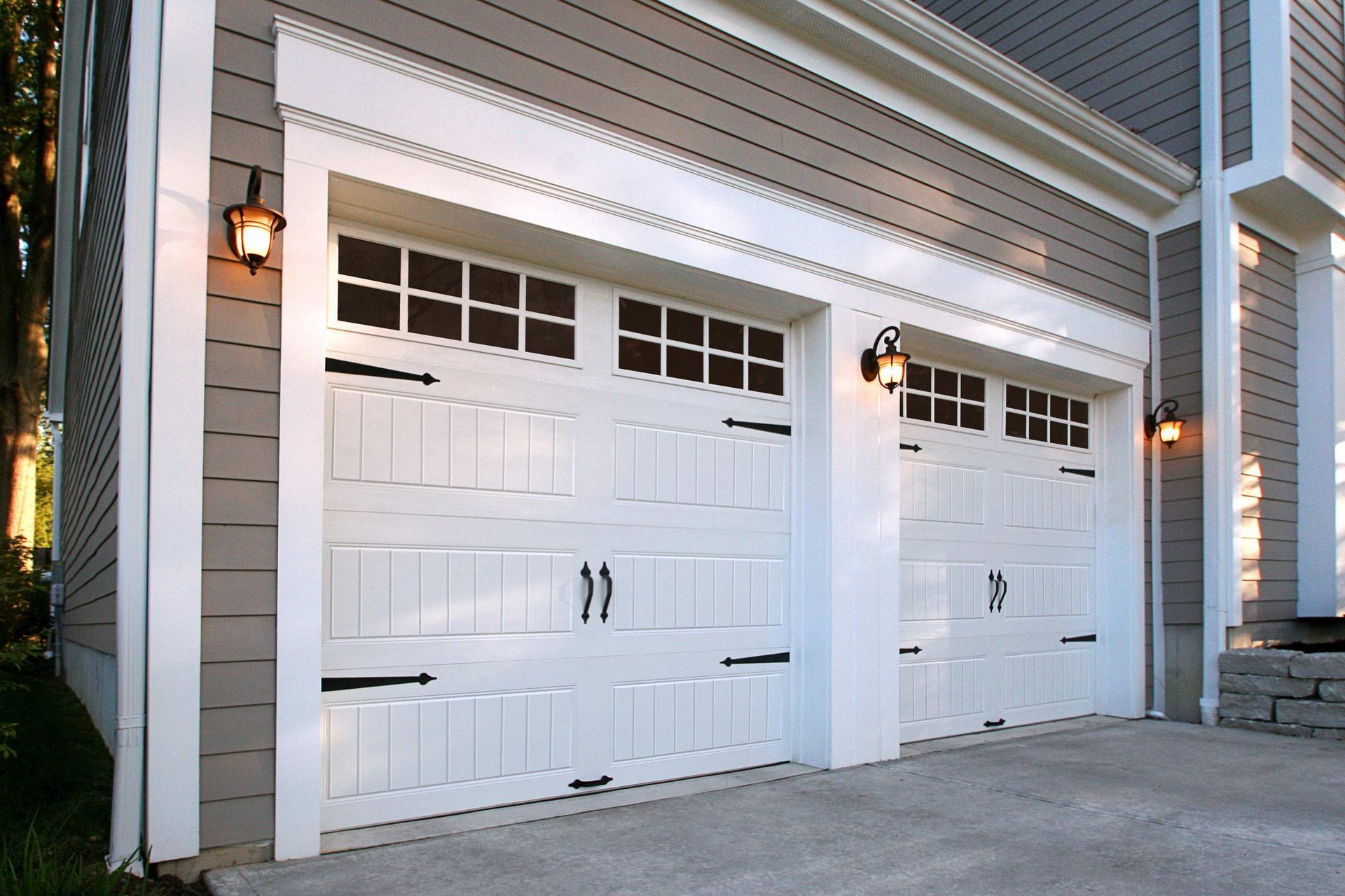 White garage doors with black hardware and windows, flanked by sconces. Gray siding.
