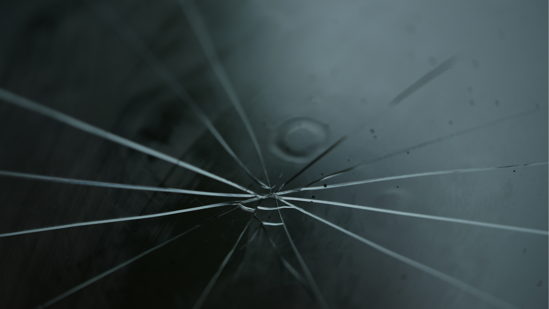 Cracked dark glass with spiderweb fractures radiating from a central impact point