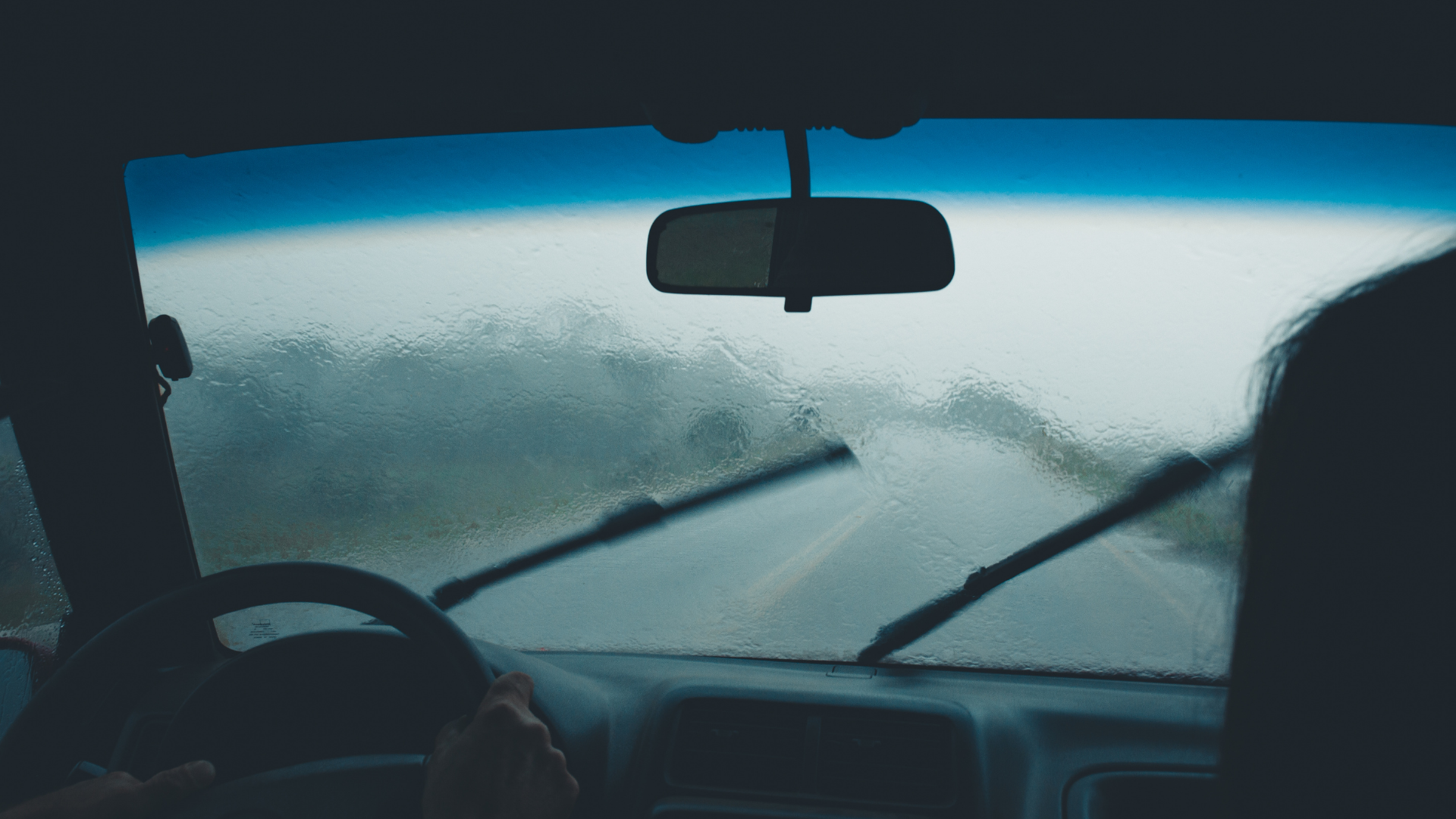 Driver’s view through a rain-soaked windshield with wipers clearing fog on a gray road
