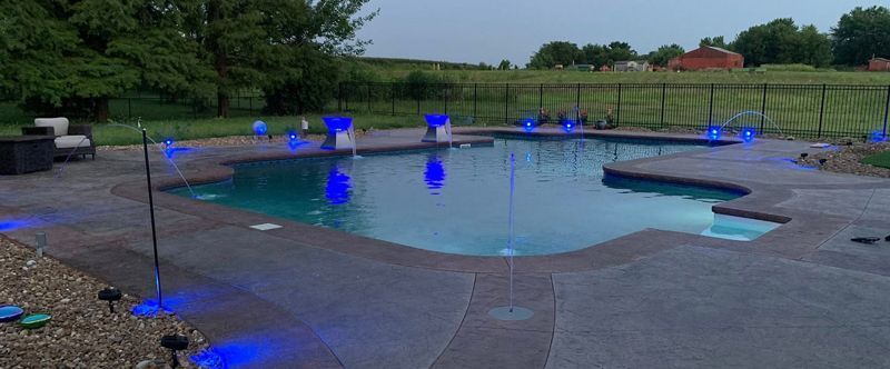 Outdoor pool with glowing blue fountains and a stone deck in a grassy park setting
