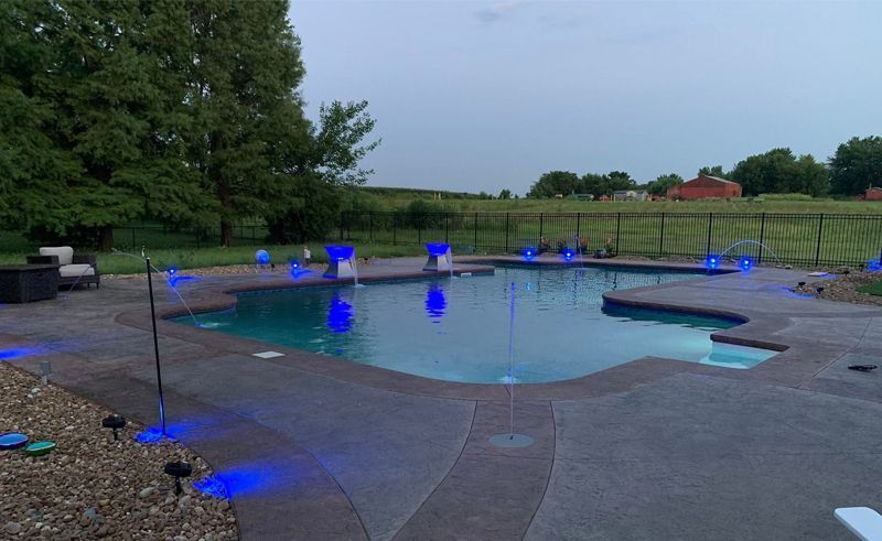 Outdoor pool with glowing blue lights beside a concrete patio and grassy field at dusk