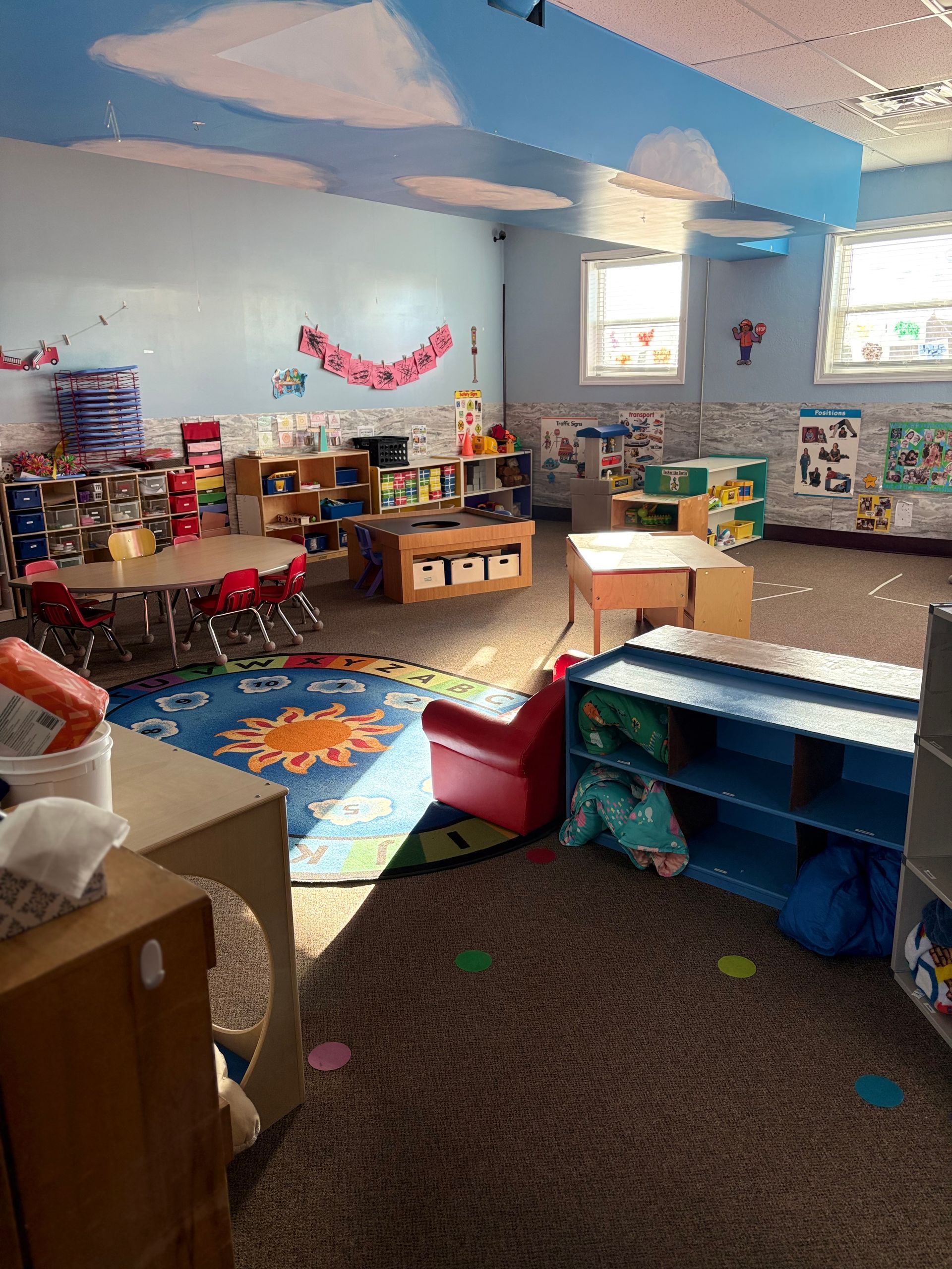 A brightly lit, colorful preschool classroom with tables, a circular rug, storage shelves, and clouds painted on the ceiling.