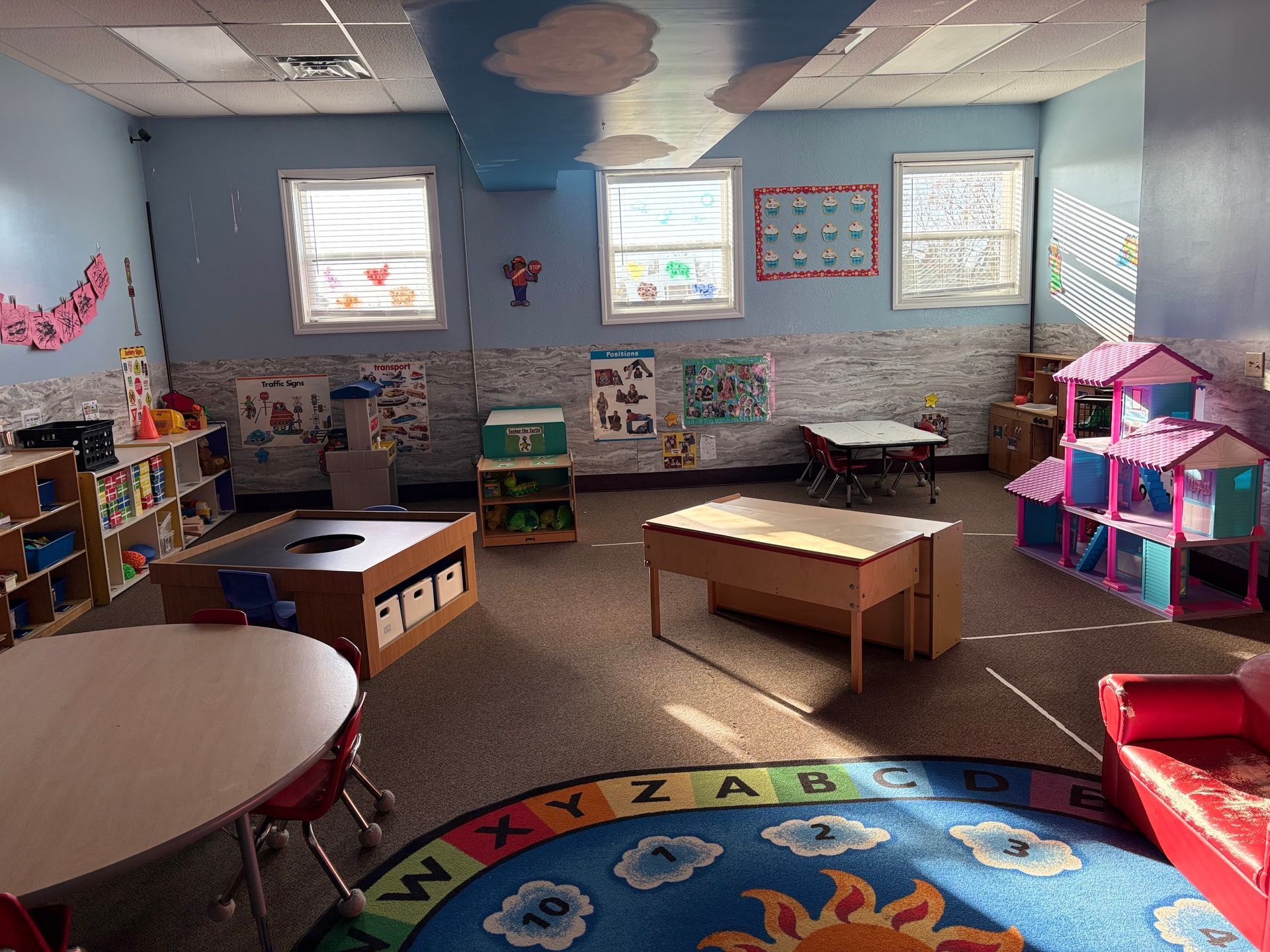 A brightly lit preschool classroom with tables, chairs, a colorful rug, a toy house, and educational wall posters.