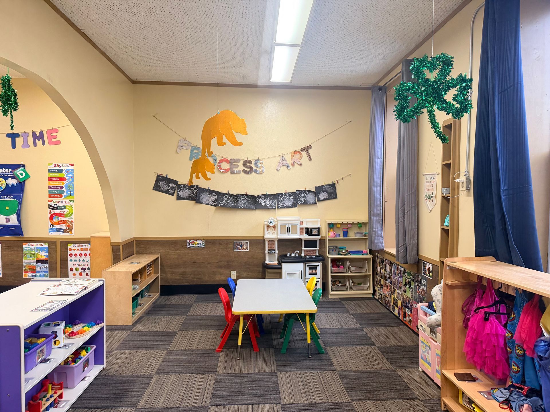 A bright preschool classroom with a central table, art display on the wall, and various shelves for toys and materials.