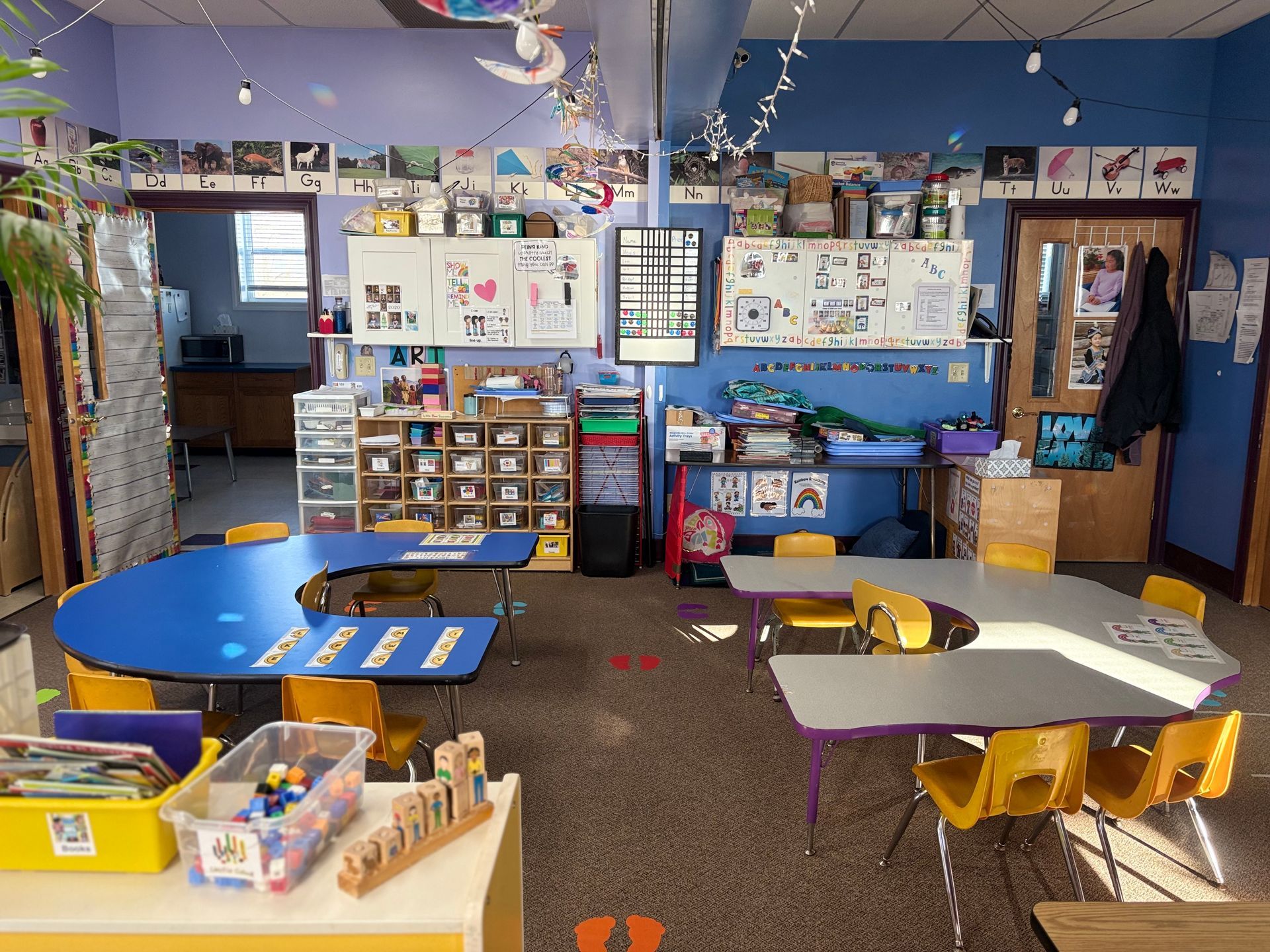 A colorful classroom with blue walls, two horseshoe-shaped tables, yellow chairs, storage shelves, and learning materials.
