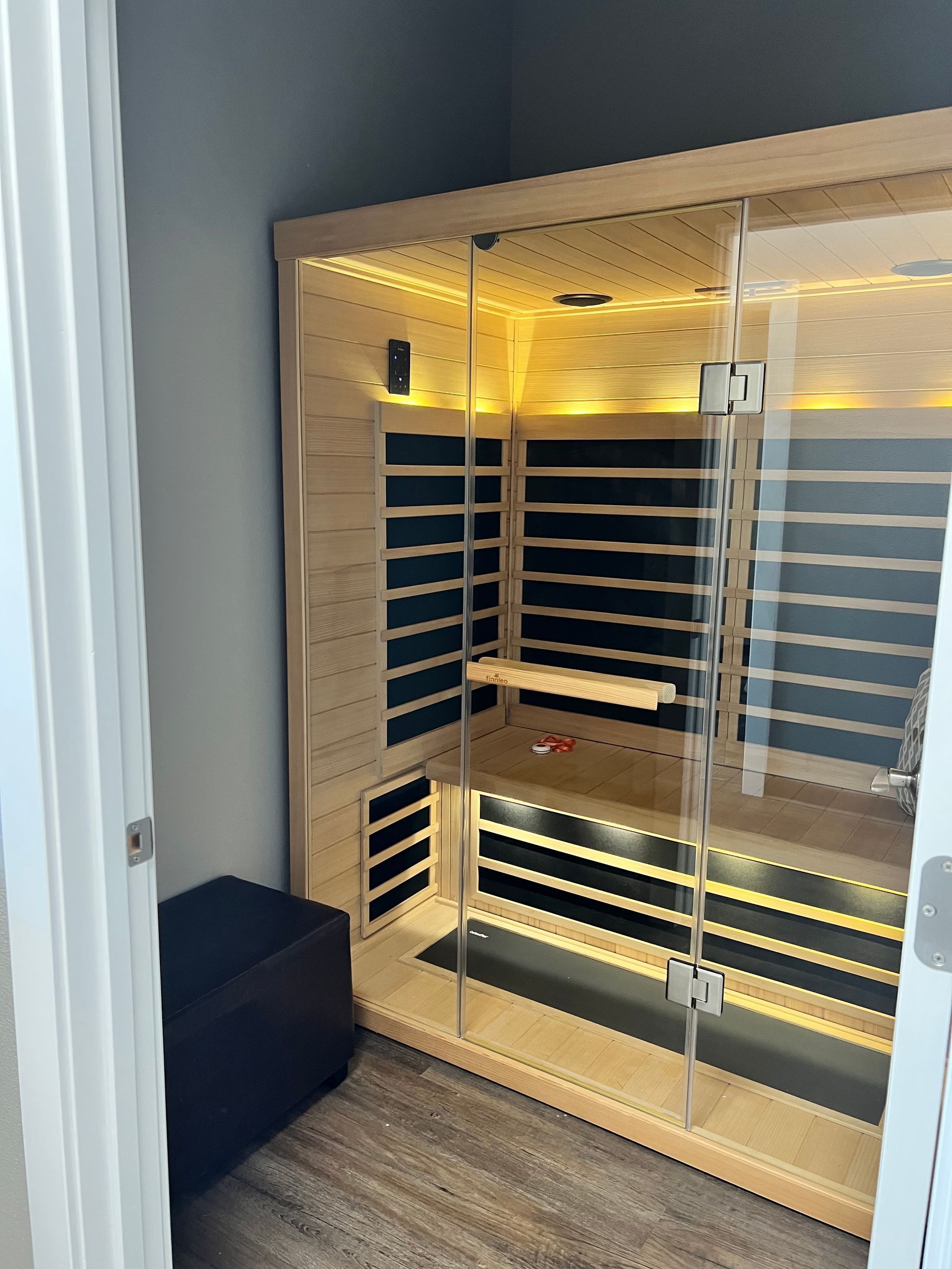 A wooden infrared sauna with glass doors, illuminated interior, and a small black bench.
