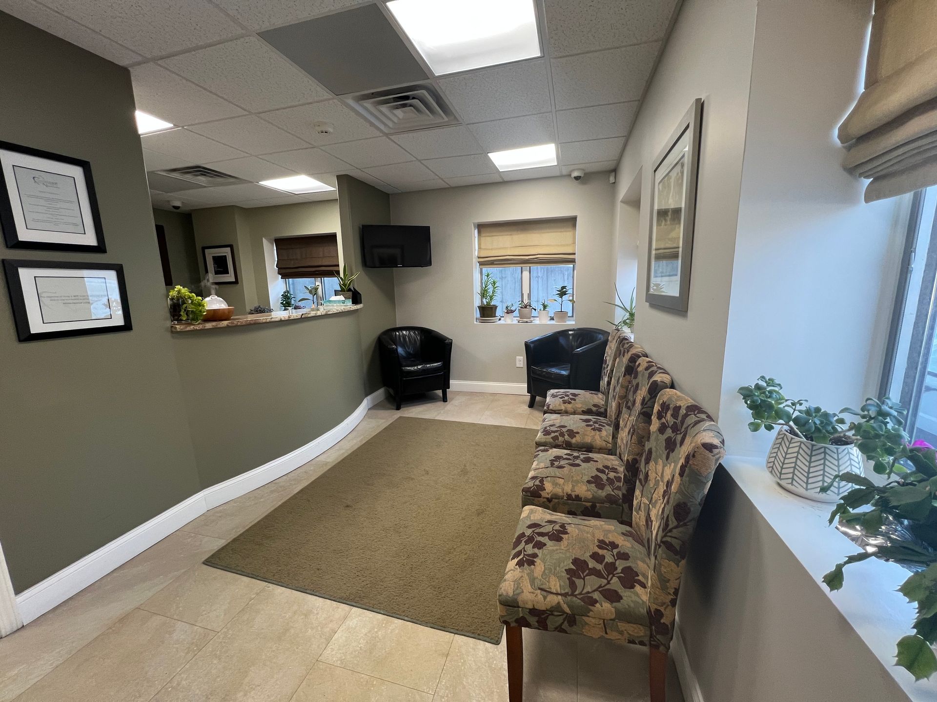 Waiting room with brown and olive green walls, two black chairs, patterned sofa, and window with blinds.