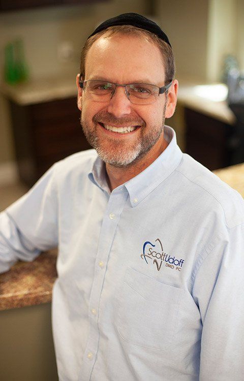Man in glasses and kippah smiling, wearing light blue shirt with logo in office setting.