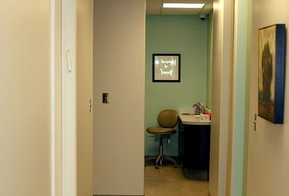 Hallway leading to an examination room with a chair, counter, and artwork. Pastel green walls, neutral doors.