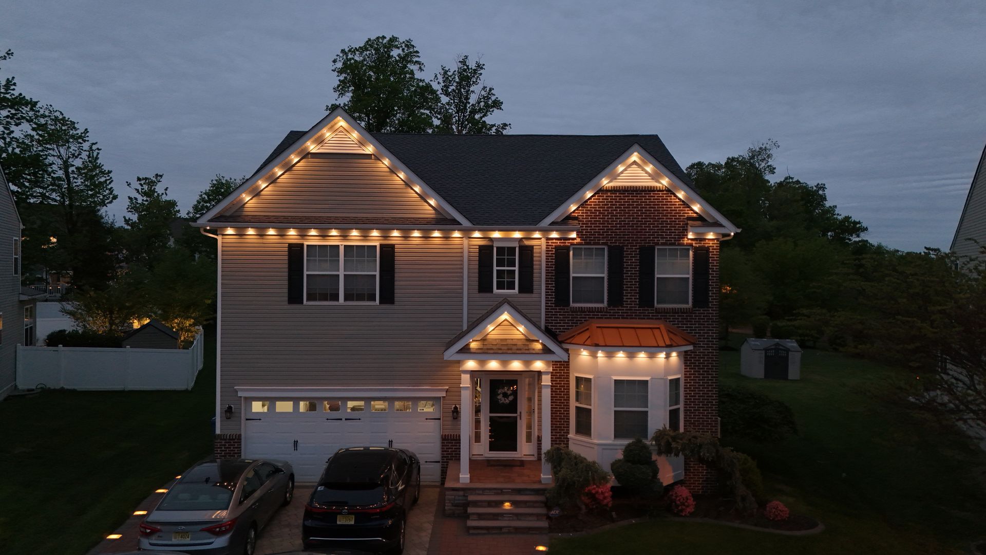 Two-story suburban house at dusk with lit roofline and front porch lights