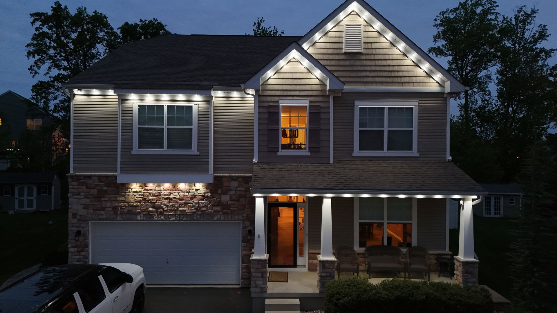 Two-story suburban house at dusk with lit windows, front porch, and garage illuminated by exterior lights
