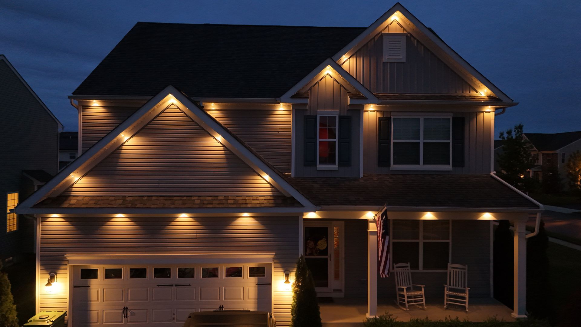 Two-story house at dusk with warm exterior lights illuminating the garage and front porch.