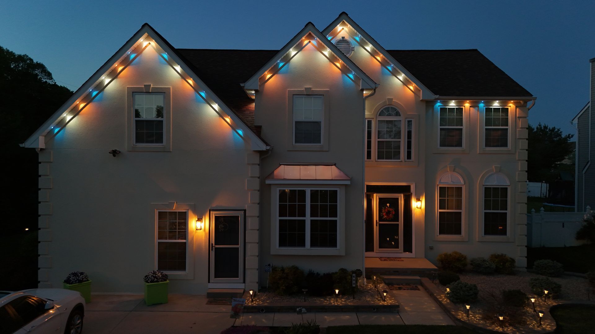Two-story house at dusk with warm porch lights and colorful roofline holiday lights