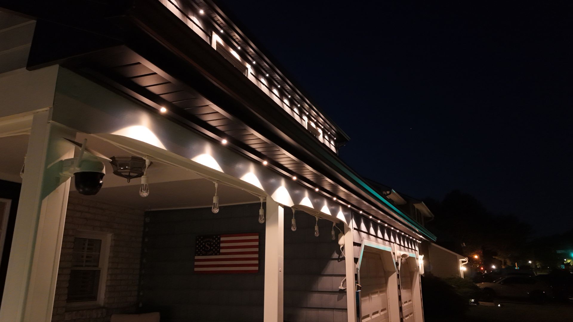 Night view of a house porch with lit eaves, hanging lanterns, and an American flag under a dark sky