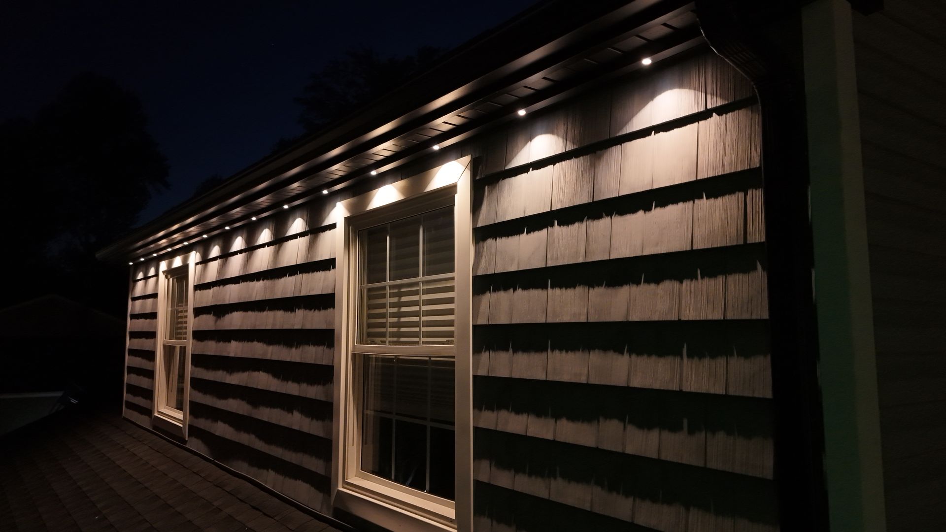 Nighttime exterior of a house with lit windows and dark horizontal siding under porch lights
