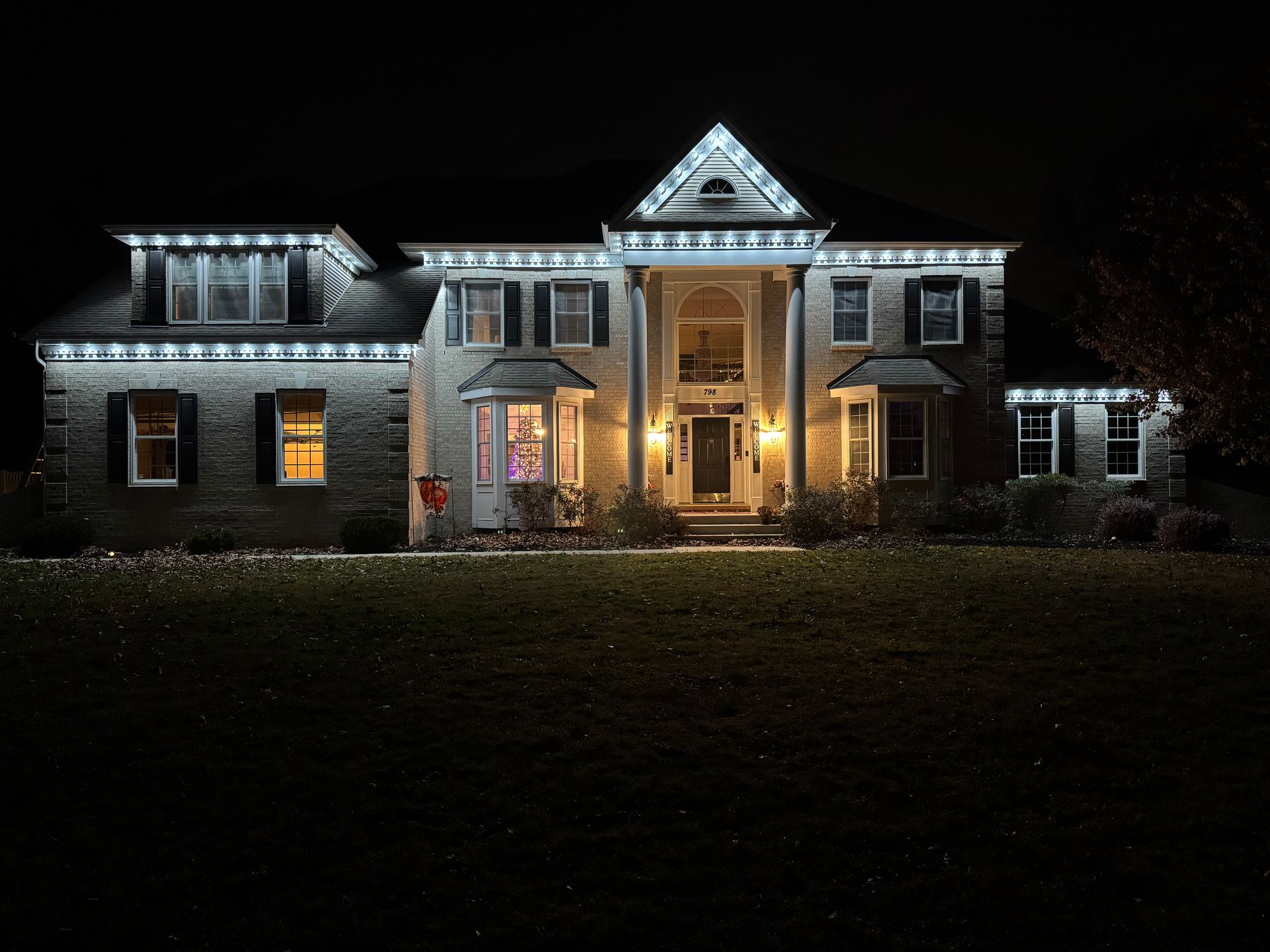 Nighttime view of a large brick house lit with white holiday lights and warm glowing windows