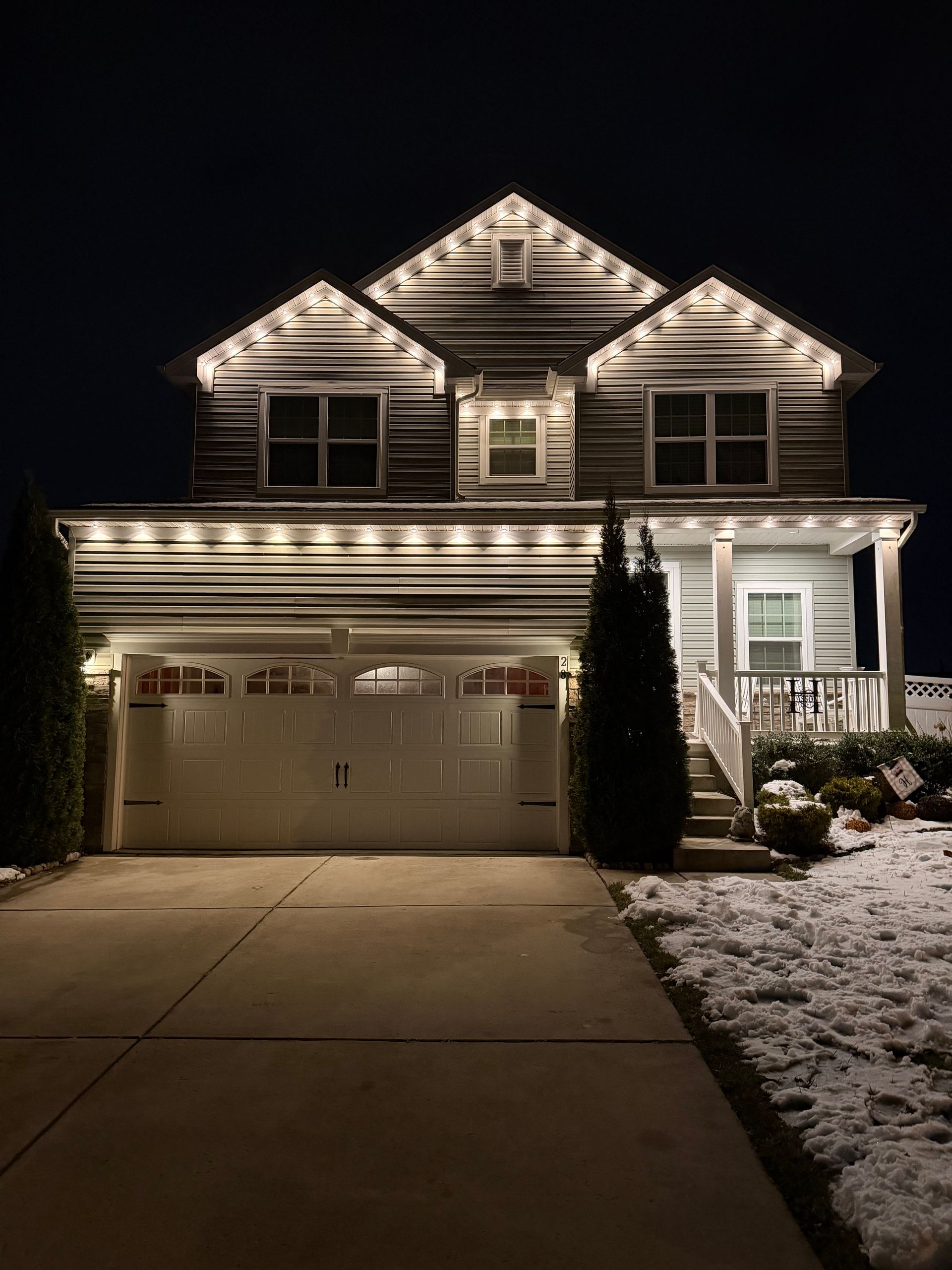 Two-story house at night with warm porch lights, lit garage, and a snow-covered lawn