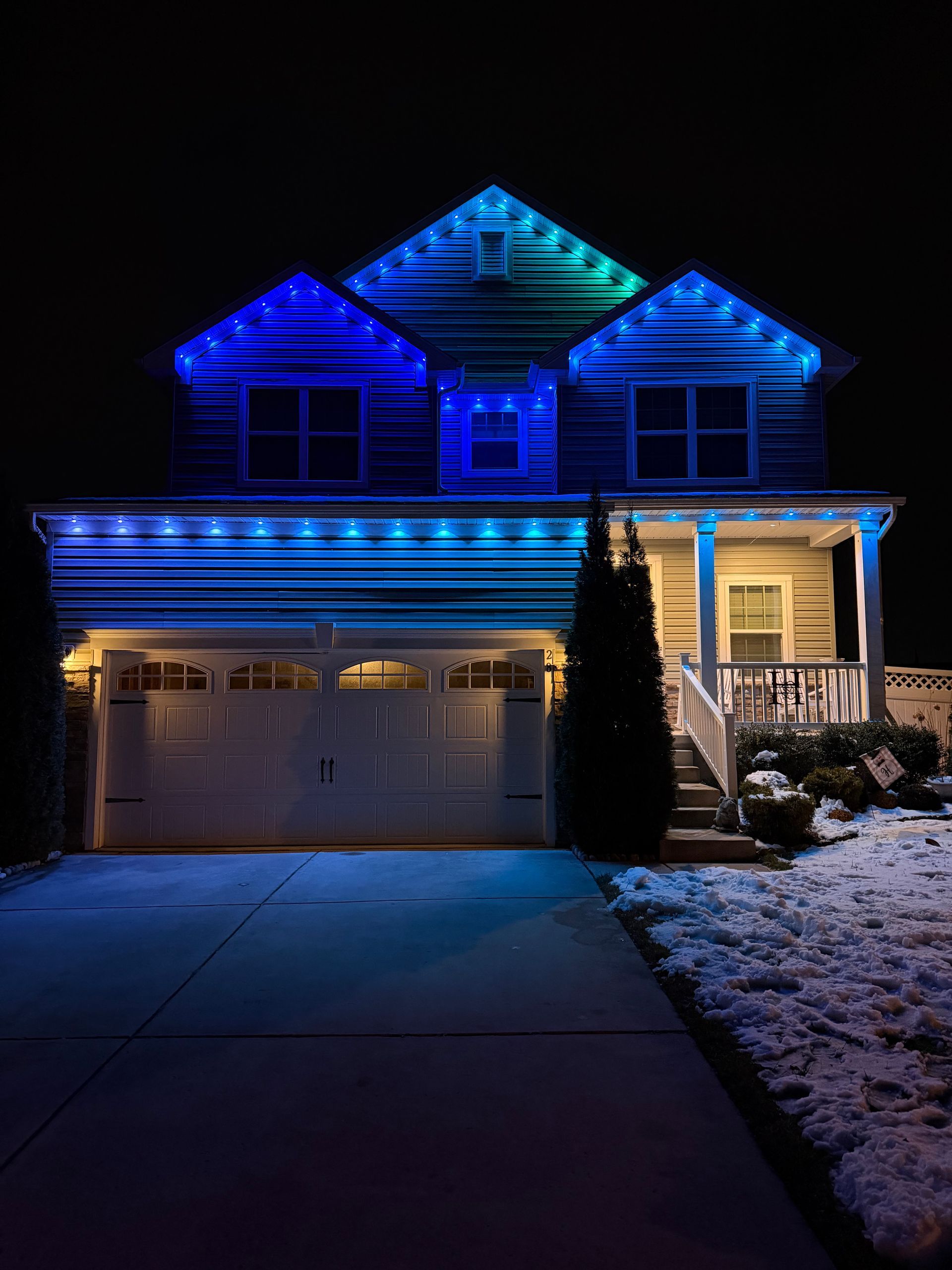 Two-story house at night lit with blue holiday lights and a glowing porch on a dark street