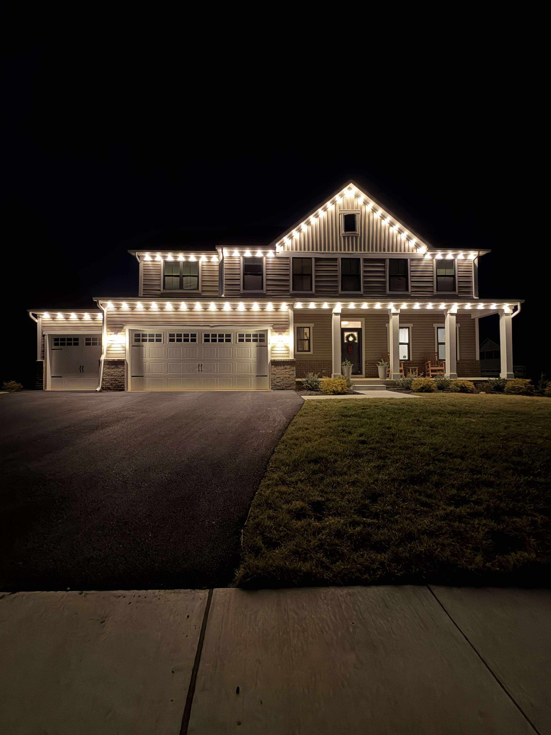 Large house at night with warm Christmas lights, viewed from the street and driveway.