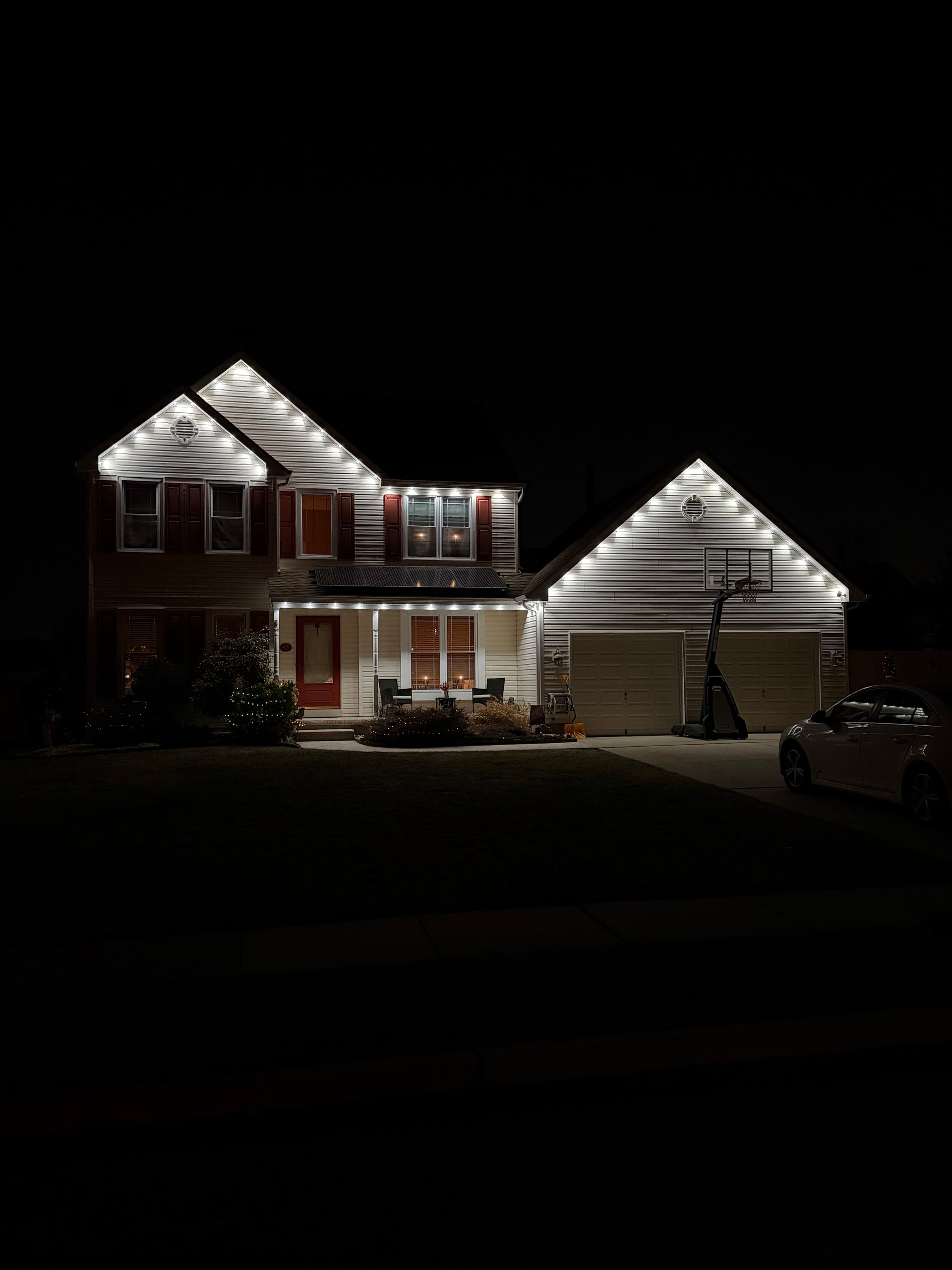 Two-story house and garage lit with white holiday lights at night, with a car parked in the driveway.